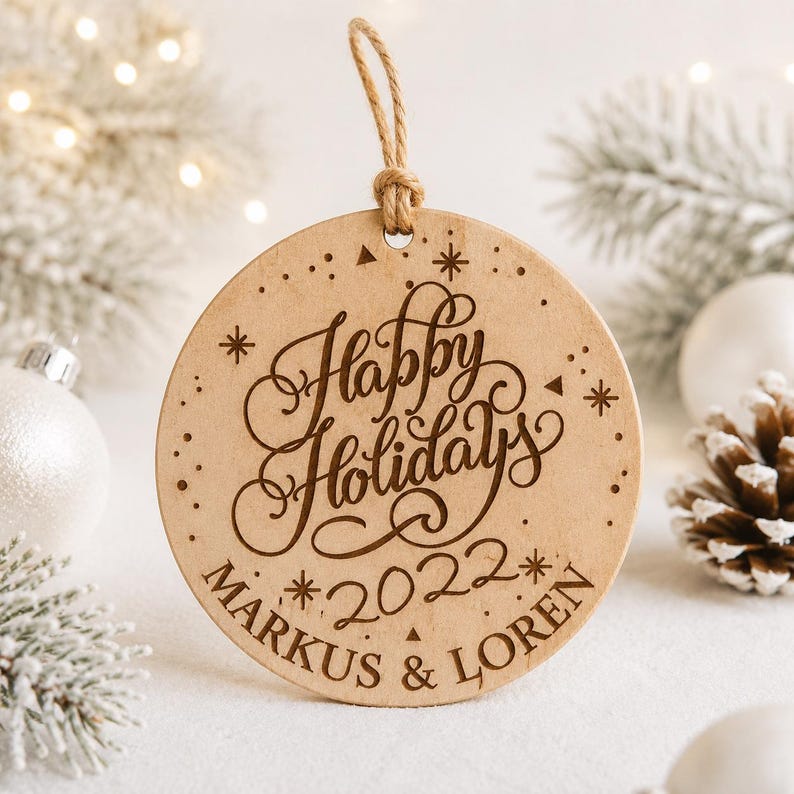 K&ouml;nnte beinhalten: Ein runder, hellfarbener Holzanh&auml;nger mit dem lasergravierten Text Happy Holidays, dem Jahr 2022 und den Namen Markus & Loren. Der Anh&auml;nger ist an einer d&uuml;nnen, naturfarbenen Juteschnur befestigt, die oben in der Mitte angebracht ist.
