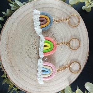 Könnte beinhalten: Drei handgefertigte Regenbogen-Schlüsselanhänger mit goldfarbener Hardware und weißem Fransen. Jeder Schlüsselanhänger hat ein anderes Farbschema: Blau, Gelb und Braun; Rosa, Grün und Gelb; und Rosa, Blau und Creme. Die Schlüsselanhänger werden auf einer Holzoberfläche präsentiert.