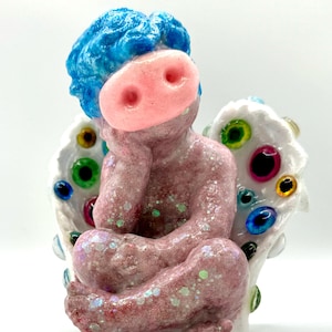 Puede incluir: Una figurita de querubín rosa con cabello azul y una nariz de cerdo se sienta en un trono blanco con ojos coloridos. La figurita está cubierta de purpurina.