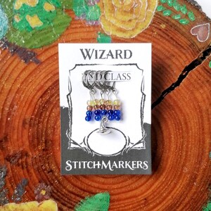May include: Set of four stitch markers with a wizard hat charm. The markers are made of silver rings with blue, yellow, and brown beads. The card reads "Wizard Class Stitch Markers".