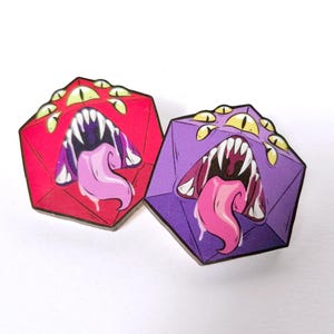 May include: Two dice with a purple and red color scheme. Each die has a cartoon monster face with multiple eyes and a large open mouth with a tongue sticking out.