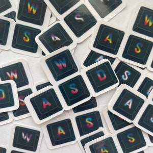 May include: A collection of square stickers featuring the letters WASD in a rainbow gradient font on a black background. The stickers are scattered across a white surface, with some overlapping. The letters are stylized to resemble keyboard keys.