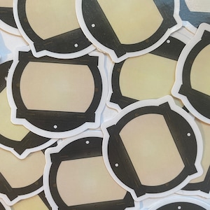 May include: A collection of round stickers with a cream-colored center and a dark brown, stylized frame. The frame has a white border and small white dots. The stickers are scattered across a surface.