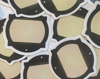 May include: A collection of round stickers with a cream-colored center and a dark brown, stylized frame. The frame has a white border and small white dots. The stickers are scattered across a surface.