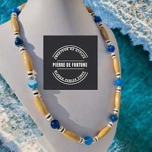 Puede incluir: Un collar de cuentas de madera y ágata azul con una cuenta de acento rayada en blanco y negro. El collar está en un maniquí blanco sobre un fondo de agua azul y blanco. La etiqueta en el maniquí dice "Createur de Styles Pierre de Fortune Bijoux Perles Fines".