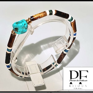 Peut inclure: Un bracelet en perles brunes et blanches avec un accent en turquoise. Le bracelet est sur un présentoir transparent. Le texte "Pierre de Fortune Bijoux Perles Fines" est visible dans le coin inférieur droit de l'image.