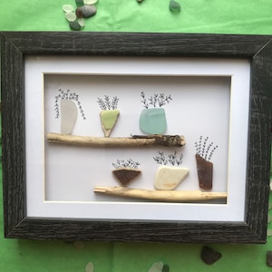 Puede incluir: Una obra de arte enmarcada que presenta una colección de piezas de vidrio de mar dispuestas para parecer plantas en maceta. El vidrio de mar es de varios colores, incluyendo blanco, verde, azul, marrón y transparente. La obra de arte está sobre un fondo blanco y enmarcada en un marco gris oscuro.