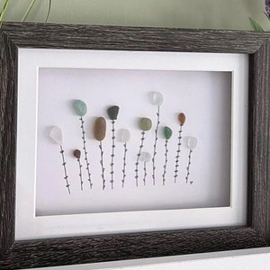 Puede incluir: Una obra de arte enmarcada que presenta una escena de la naturaleza con una variedad de piedras de colores dispuestas para parecer flores. Las piedras son de tonos verdes, marrones y blancos, y están dispuestas sobre un fondo blanco. La obra de arte está enmarcada en un marco de madera gris oscuro.