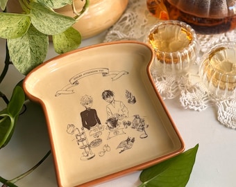 May include: A beige ceramic plate shaped like a slice of bread with a brown rim. The center features a black line drawing of two people preparing tea, with a banner above reading "ATELIER AFTERNOON TEA" and various tea-related items.