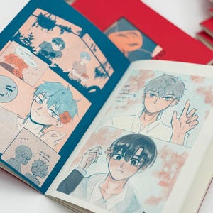 May include: Open book with manga-style illustrations. The pages display colorful panels with male characters, floral accents, and speech bubbles. The book has a red cover and is surrounded by other books.
