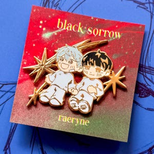 May include: Enamel pin featuring two cartoon figures in white clothing, holding hands, with gold star accents. The pin is on a red card with the text "black sorrow" and "raeryne".