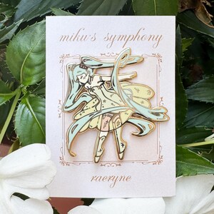 May include: A gold-colored enamel pin featuring a stylized anime character with long, flowing teal hair and a light yellow dress. The pin is on a white card with the text "miku's symphony" and "raeryne". The background includes green leaves and white flowers.
