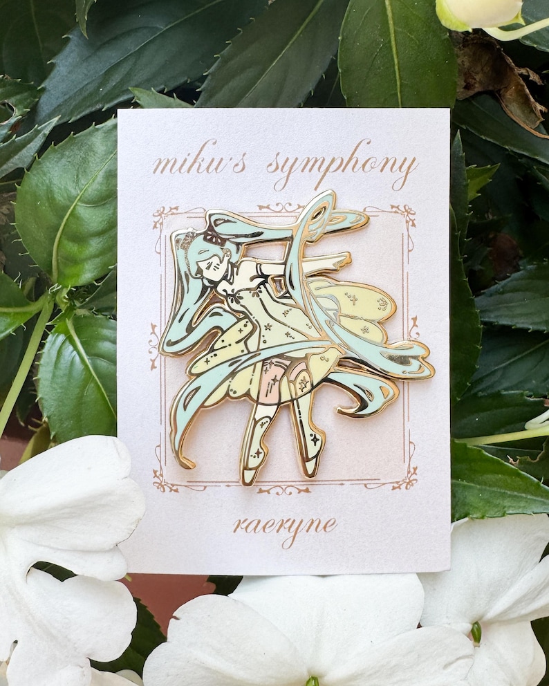May include: A gold-colored enamel pin featuring a stylized anime character with long, flowing teal hair and a light yellow dress. The pin is on a white card with the text "miku's symphony" and "raeryne". The background includes green leaves and white flowers.
