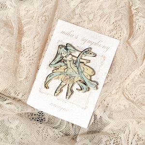 May include: A gold-colored enamel pin featuring a stylized anime character with long, flowing teal hair and a yellow dress. The pin is on a white card with the text "miku's symphony" and the name "raeryne".