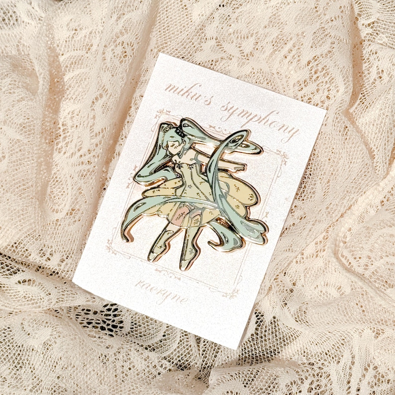 May include: A gold-colored enamel pin featuring a stylized anime character with long, flowing teal hair and a yellow dress. The pin is on a white card with the text "miku's symphony" and the name "raeryne".