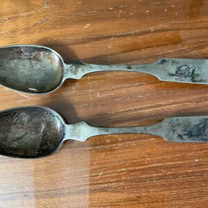 May include: Two antique silver spoons with ornate handles. The spoons have a tarnished finish and are engraved with the initials "B.L."
