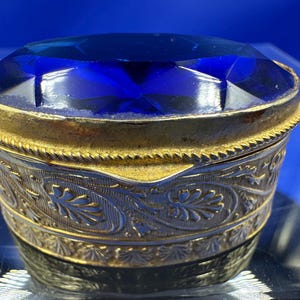 May include: A small, round, gold-colored trinket box with an ornate design. The lid is made of a large, faceted, blue glass gem.