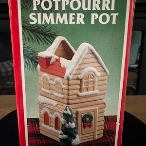 May include: A ceramic house-shaped potpourri simmer pot with a red roof, white snow accents, and a green Christmas tree. The box reads "Potpourri Simmer Pot".