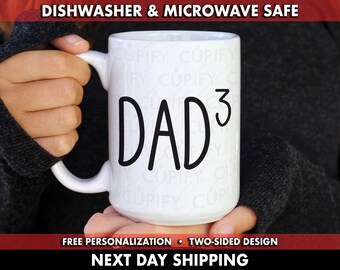 Op de afbeelding: Een witte keramische mok met zwarte tekst die "DAD^3" luidt. De mok wordt vastgehouden door een hand die een zwarte trui draagt. De tekst "DISHWASHER & MICROWAVE SAFE" staat bovenaan de afbeelding. De tekst "FREE PERSONALIZATION - TWO-SIDED DESIGN" staat onderaan de afbeelding. De tekst "NEXT DAY SHIPPING" staat onderaan de afbeelding.