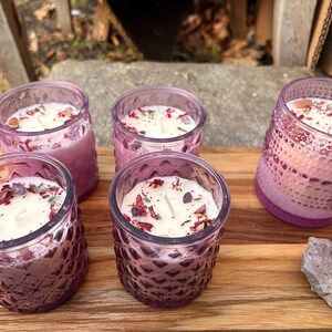 May include: Five purple glass candles with white wax, decorated with dried flowers and crystals, are arranged on a wooden surface. The candles have a textured design. The candles are a light purple colour.
