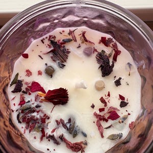May include: A lit candle in a clear glass jar. The candle has a creamy white wax surface, decorated with dried red petals, lavender, and small stones. The jar has a textured, patterned design.
