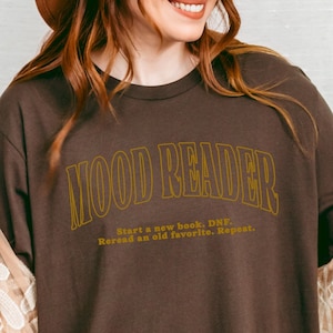 Könnte beinhalten: Ein braunes T-Shirt mit dem Text "Mood Reader" in goldenen Buchstaben. Der Text darunter lautet "Start a new book. DNF. Reread an old favorite. Repeat."
