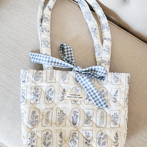 Puede incluir: Bolso tote acolchado con fondo blanco e ilustraciones florales azules. El bolso tiene asas a juego y un lazo de cuadros azul. El texto "fearfully and wonderfully made" está en una pequeña etiqueta.