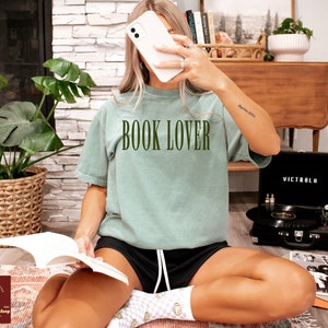 Op de afbeelding: Een vrouw draagt een lichtgroen T-shirt met de tekst "BOOK LOVER" in donkergroene letters. Ze zit op een tapijt met een boek op haar schoot en een platenspeler op de achtergrond.