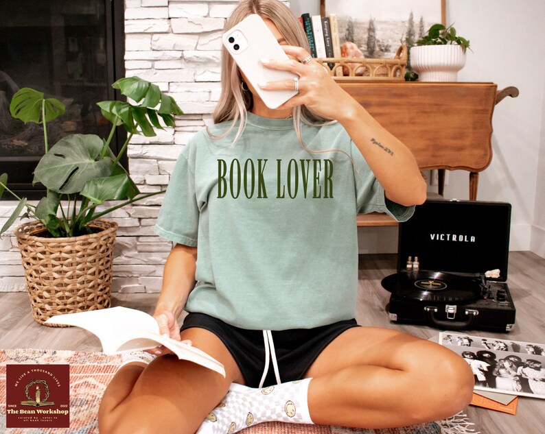 Op de afbeelding: Een vrouw draagt een lichtgroen T-shirt met de tekst "BOOK LOVER" in donkergroene letters. Ze zit op een tapijt met een boek op haar schoot en een platenspeler op de achtergrond.