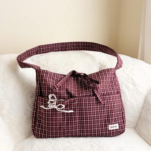 May include: A burgundy and white checked tote bag with a single shoulder strap. The bag features a front pocket with a white decorative bow and a small white tag that says "BOOK BAG".