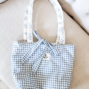 Puede incluir: Un bolso tote de cuadros vichy en azul y blanco con un cierre de lazo en la parte superior. El bolso cuenta con dos asas acolchadas sobre una base de tela blanca con un patr&oacute;n floral azul. Una peque&ntilde;a etiqueta rectangular blanca dice THE BEAN WORKSHOP.