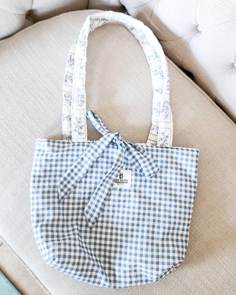 Puede incluir: Un bolso tote de cuadros vichy en azul y blanco con un cierre de lazo en la parte superior. El bolso cuenta con dos asas acolchadas sobre una base de tela blanca con un patr&oacute;n floral azul. Una peque&ntilde;a etiqueta rectangular blanca dice THE BEAN WORKSHOP.