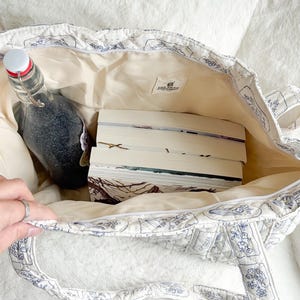 Puede incluir: Un bolso tote acolchado con base blanca y patr&oacute;n de l&iacute;neas florales azules, que contiene tres libros apilados y una botella de vidrio con tapa blanca. Una peque&ntilde;a etiqueta rectangular en el interior muestra el texto The Book Workshop Est 2024.