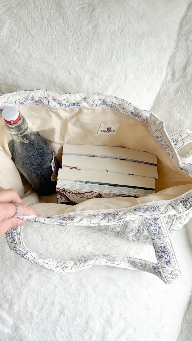 Puede incluir: Un bolso tote acolchado con base blanca y patr&oacute;n de l&iacute;neas florales azules, que contiene tres libros apilados y una botella de vidrio con tapa blanca. Una peque&ntilde;a etiqueta rectangular en el interior muestra el texto The Book Workshop Est 2024.