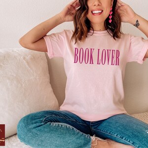 Op de afbeelding: Een roze T-shirt met de tekst "BOOK LOVER" in roze op de voorkant. Het shirt wordt gedragen door een persoon die op een witte en cr&egrave;mekleurige bank zit.