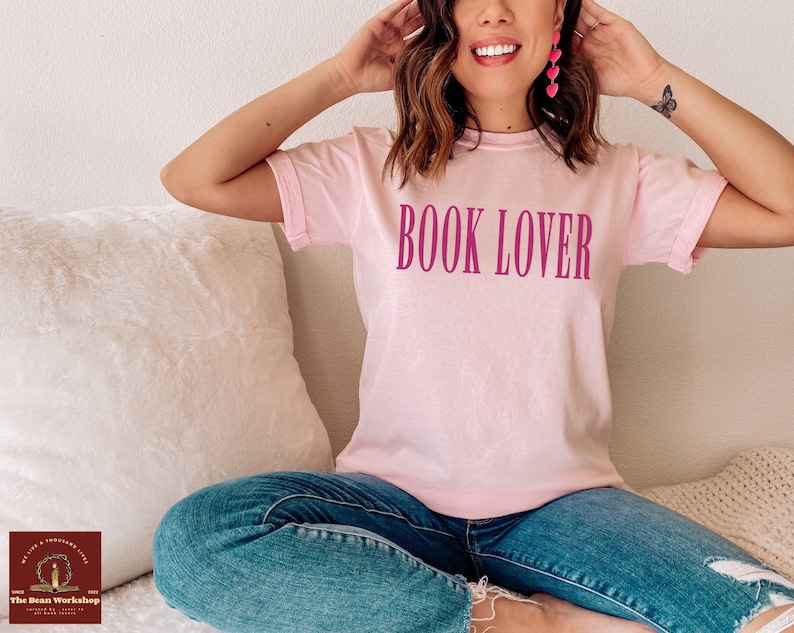 Op de afbeelding: Een roze T-shirt met de tekst "BOOK LOVER" in roze op de voorkant. Het shirt wordt gedragen door een persoon die op een witte en cr&egrave;mekleurige bank zit.