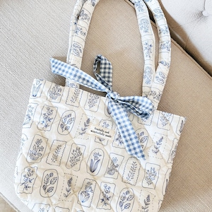 Puede incluir: Un bolso tote acolchado en color blanco roto con un estampado bot&aacute;nico azul de varias flores en marcos rectangulares. El bolso tiene dos asas acolchadas a juego y una cinta de cuadros vichy azul y blanca atada en la parte superior. Una etiqueta dice fearfully and wonderfully made.