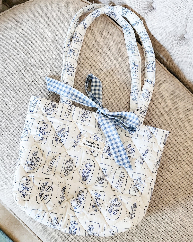 Puede incluir: Un bolso tote acolchado en color blanco roto con un estampado bot&aacute;nico azul de varias flores en marcos rectangulares. El bolso tiene dos asas acolchadas a juego y una cinta de cuadros vichy azul y blanca atada en la parte superior. Una etiqueta dice fearfully and wonderfully made.