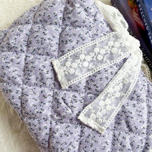 May include: A quilted lavender fabric pouch featuring a small white floral pattern. The pouch is accented with a decorative white lace ribbon trim that has an embroidered floral design, adding texture to the soft, padded surface of the item.