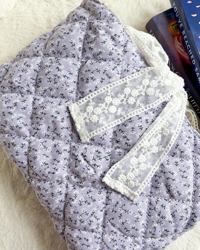 May include: A quilted lavender fabric pouch featuring a small white floral pattern. The pouch is accented with a decorative white lace ribbon trim that has an embroidered floral design, adding texture to the soft, padded surface of the item.