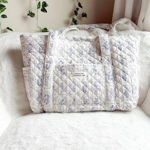 Puede incluir: Un bolso tote acolchado en color blanco roto con un estampado bot&aacute;nico azul repetido dentro de marcos rectangulares. El bolso tiene dos asas superiores, bolsillos laterales y un bolsillo frontal central con una etiqueta que dice fearfully and wonderfully made.