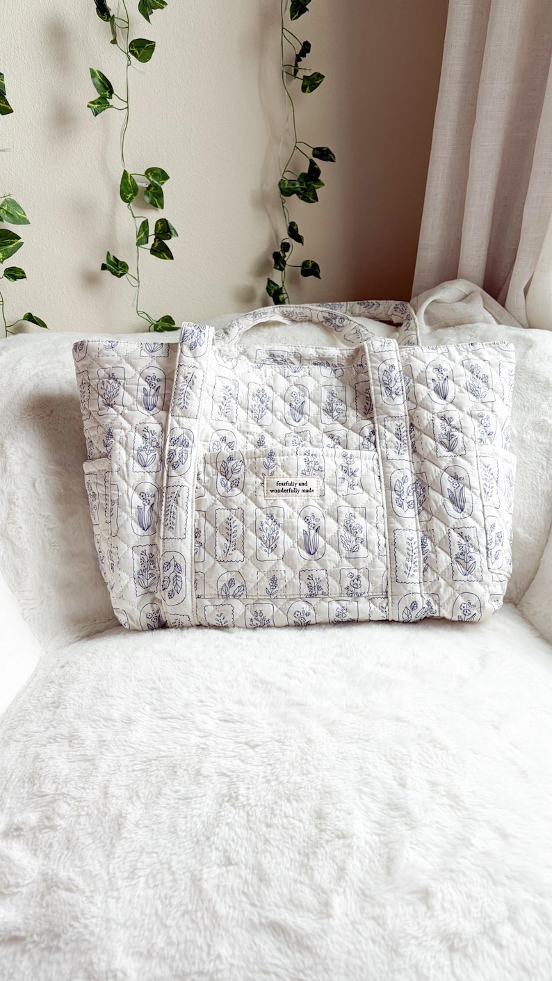 Puede incluir: Un bolso tote acolchado en color blanco roto con un estampado bot&aacute;nico azul repetido dentro de marcos rectangulares. El bolso tiene dos asas superiores, bolsillos laterales y un bolsillo frontal central con una etiqueta que dice fearfully and wonderfully made.