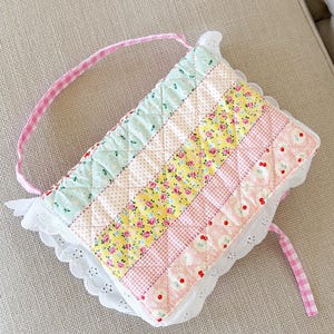 May include: A quilted rectangular pouch featuring diagonal stripes of floral and gingham fabric in pastel shades of mint green, yellow, and pink. The pouch is trimmed with white eyelet lace and includes a pink and white gingham fabric handle.