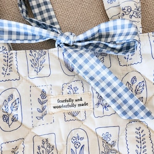 Puede incluir: Un bolso tote acolchado de tela blanca con un patr&oacute;n bot&aacute;nico de l&iacute;neas azules y un lazo de cuadros vichy azul y blanco. Una etiqueta rectangular de tela est&aacute; cosida en el centro con el texto, fearfully and wonderfully made.