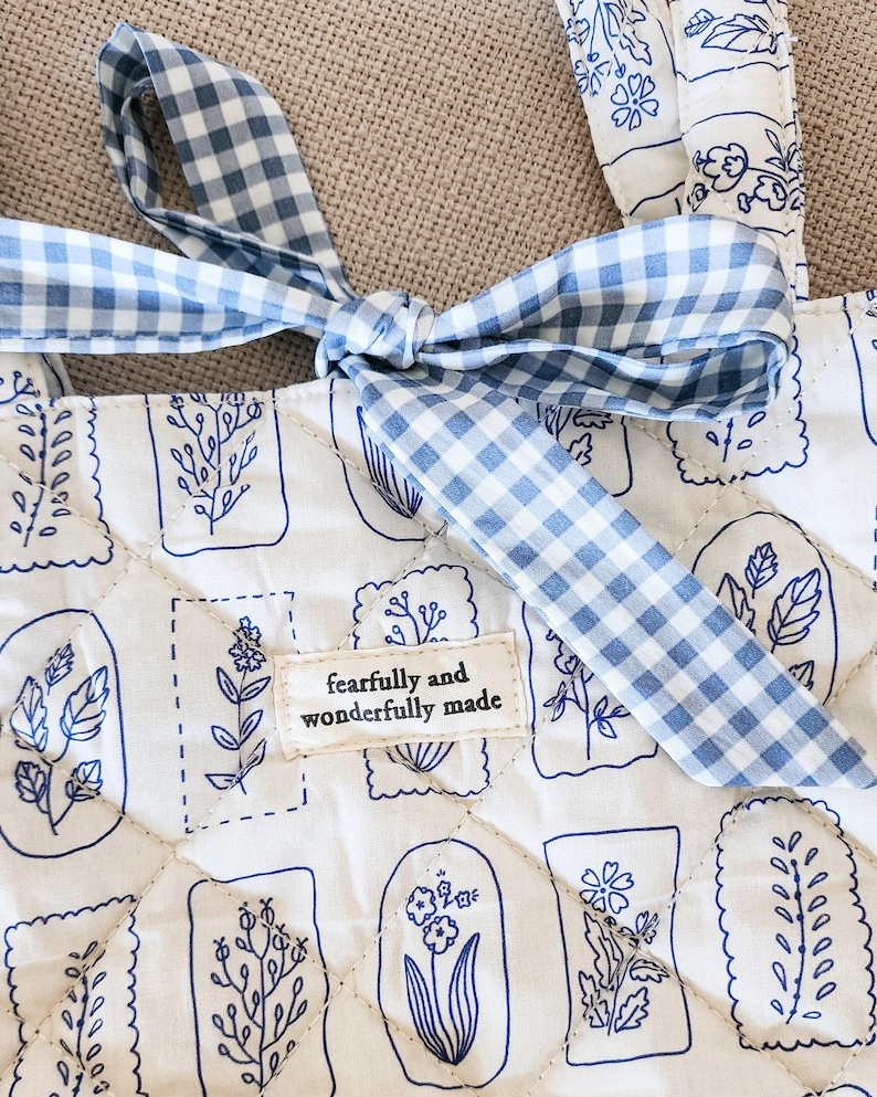 Puede incluir: Un bolso tote acolchado de tela blanca con un patr&oacute;n bot&aacute;nico de l&iacute;neas azules y un lazo de cuadros vichy azul y blanco. Una etiqueta rectangular de tela est&aacute; cosida en el centro con el texto, fearfully and wonderfully made.