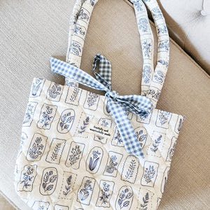 Puede incluir: Un bolso tote acolchado en color blanco roto con un patr&oacute;n de sellos bot&aacute;nicos azules y dos asas a juego. Una cinta de cuadros vichy azul y blanca est&aacute; atada en la base de una de las asas. Una peque&ntilde;a etiqueta rectangular dice "fearfully and wonderfully made".