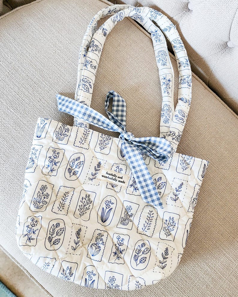 Puede incluir: Un bolso tote acolchado en color blanco roto con un patr&oacute;n de sellos bot&aacute;nicos azules y dos asas a juego. Una cinta de cuadros vichy azul y blanca est&aacute; atada en la base de una de las asas. Una peque&ntilde;a etiqueta rectangular dice "fearfully and wonderfully made".
