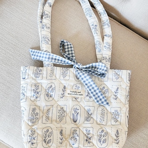 Puede incluir: Un bolso tote acolchado con base blanca y dibujos lineales de flores azules en marcos rectangulares. El bolso tiene dos asas acolchadas a juego y un lazo de cinta de cuadros vichy azul y blanco. Una etiqueta de tela dice fearfully and wonderfully made.