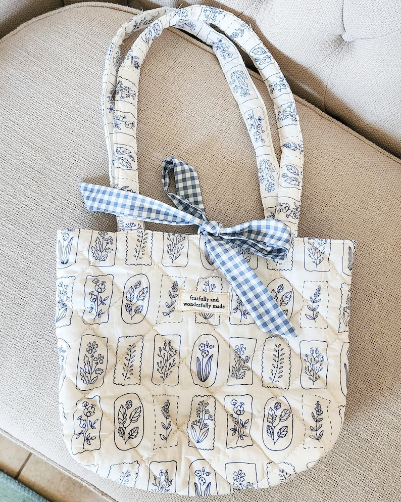 Puede incluir: Un bolso tote acolchado con base blanca y dibujos lineales de flores azules en marcos rectangulares. El bolso tiene dos asas acolchadas a juego y un lazo de cinta de cuadros vichy azul y blanco. Una etiqueta de tela dice fearfully and wonderfully made.