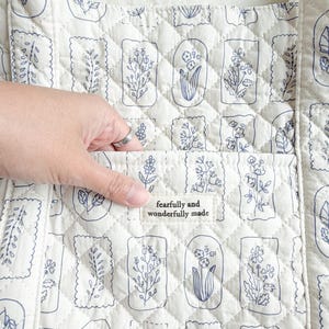 Puede incluir: Un bolso tote acolchado de color blanco roto con un patr&oacute;n repetitivo de l&iacute;neas azules de varias flores y hojas en marcos rectangulares. Una etiqueta de tela en el bolsillo frontal muestra el texto "fearfully and wonderfully made" en negro.